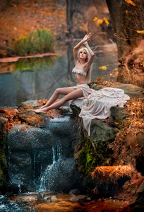 美丽的年轻女子坐在魔法森林的河流附近。的金发女郎，有着白色的衣服在秋天公园构成挑衅。童话的女孩看起来在秋天风景。浪漫的女人湖