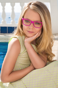 年轻漂亮的孩子女孩模型金黄色长卷发粉色眼镜和别致的绿色长礼服粉红色玻璃在池与栏杆和西班牙，希腊，圣托里尼岛海微笑