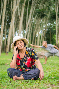 一位在花园里用手机的亚洲老妇人，一位男子在后面摆姿势