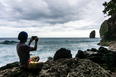 年轻人坐在岩石上和波在智能手机上的照片制作