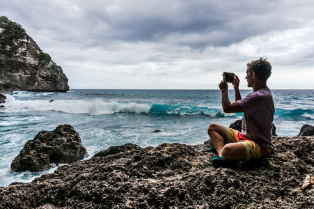 年轻人坐在岩石上和波在智能手机上的照片制作