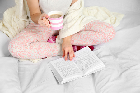女人穿着睡衣读一本书，在她的床上喝咖啡