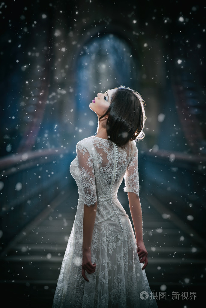 天空光和雪花落在她的脸上在长婚纱冬季风光一座桥上摆姿势的黑发女孩