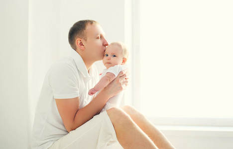 快乐年轻的父亲，在白色的房间里，靠近窗亲吻宝宝在家里