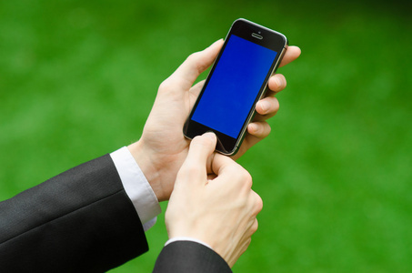 通信和商务主题 携手一套黑色的西装，在绿草的背景举行现代电话与蓝色屏幕