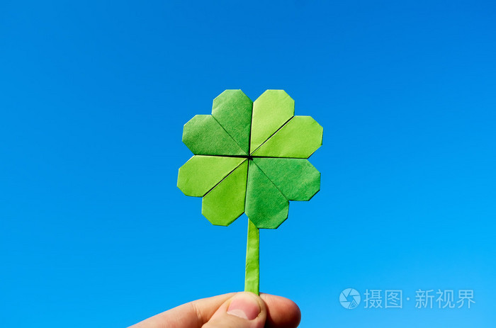 hand holding green paper origami folded shamrock on blue sky bac