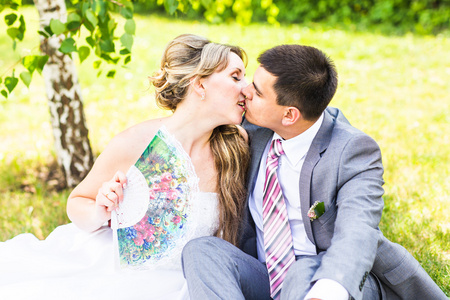 美丽的新娘和新郎坐在草和接吻。年轻的新婚夫妇