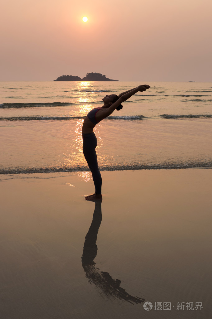年轻苗条的女人练习在日落时从海滩上瑜伽姿势