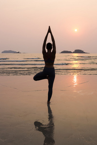 年轻苗条的女人练习在日落时从海滩上瑜伽姿势
