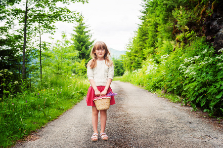 可爱的孩子女孩的 67 岁徒步旅行在森林里，抱着小篮子鲜花，穿着米色套衫和红色的裙子