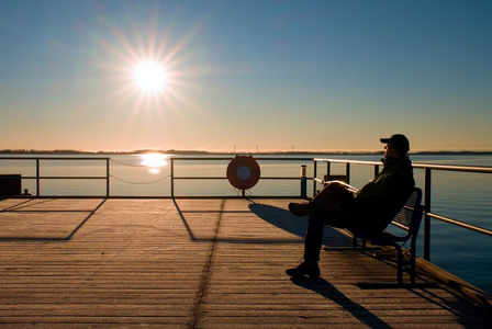 男人坐在长椅上码头建设，看着大海。晴朗的蓝天底下，光滑的水平