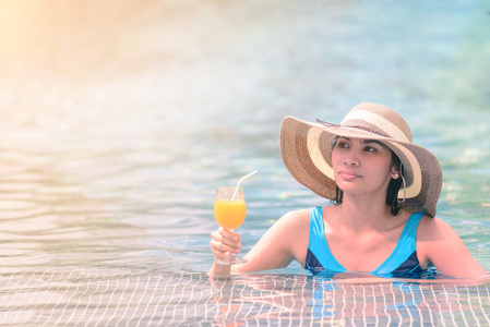 美丽的女人，戴着帽子在池畔举橙汁杯子夏天