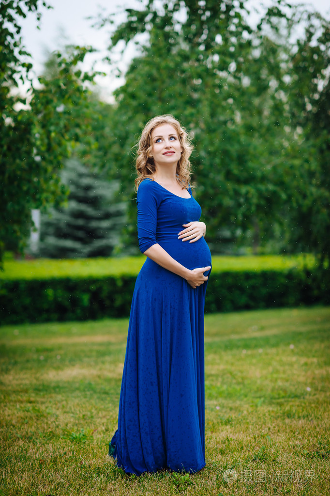 可爱年轻孕妇的抱着她的肚子长长的金色卷发的蓝色礼服微笑看着天空中