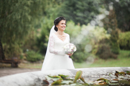 美丽的黑发新娘在优雅的白色连衣裙，抱着花束摆整齐的树木