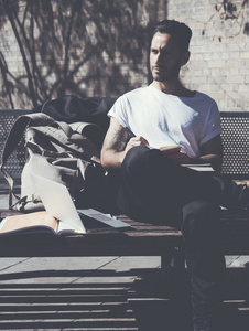 照片胡子的男人，穿着白色 t 恤，坐在城市公园，并绘制记事本。就读大学，从事的项目。书，通用设计笔记本电脑背包板凳。垂直