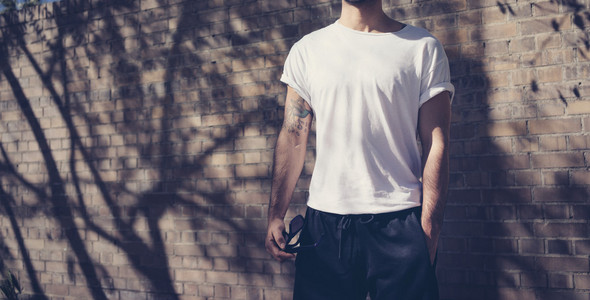 照片严重的年轻人戴着空白的白色 t 恤的纹身。站在一堵墙前面。城市街道背景。水平的样机