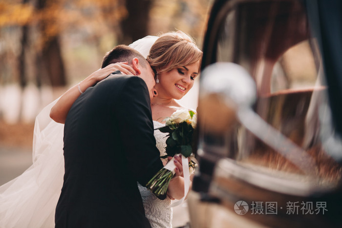 时尚的婚礼夫妇新娘新郎亲吻和拥抱附近复古车在秋天