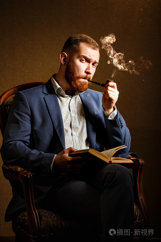 英俊的年轻男子吸烟管和读的书的蓝色西装的肖像