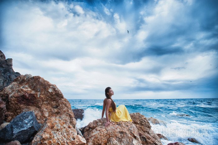 一个女孩坐在海滩上的岩石上靠着天空和大海