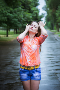 年轻漂亮的金发白种人女孩穿着短裤和衬衫在公园散步时在夏天暖雨中赤脚通过水坑。有乐趣和享受。雨落。雨滴。弄湿了衣服，把头发弄湿