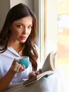年轻女子在家里坐在靠近窗口轻松的在她的客厅里看书 喝咖啡或茶