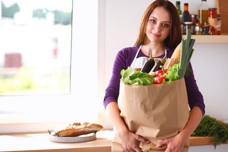 年轻女子捧着与蔬菜站在厨房里的食品杂货店购物袋
