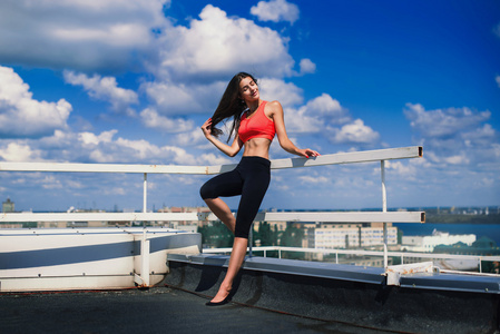画像的金发坚强的女人，在做伸展运动以上大城市的摩天大楼的屋顶上，在清晨的运动服。女性运动和性感肌肉发达的身体。健身概念