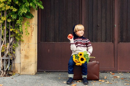 秋天的可爱的小金发男孩 4 岁，穿着暖和的套衫与引擎盖 深色牛仔布牛仔裤和蓝色的鞋子，拿着一个苹果和黄色的向日葵花束的肖像