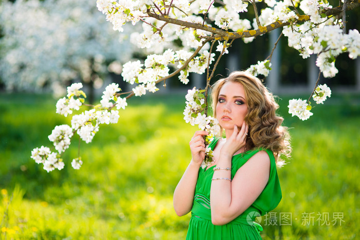 站在花朵盛开的苹果树旁边的漂亮年轻的金发女郎
