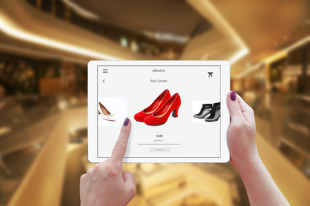 女人在线购物的平板电脑。夹持装置和选择红色的鞋子。购物中心在背景中