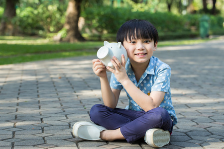 亚洲的小男孩是坐和公园里拿着他的存钱罐