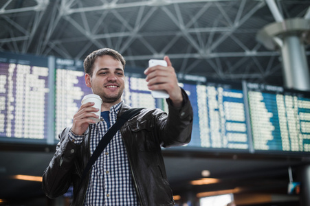 微笑在机场一杯咖啡的年轻旅行者人使时间表板前拍照