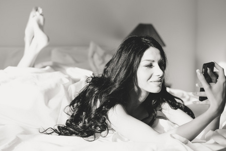调情的年轻女子躺在床上使用智能手机，以自拍照的肖像