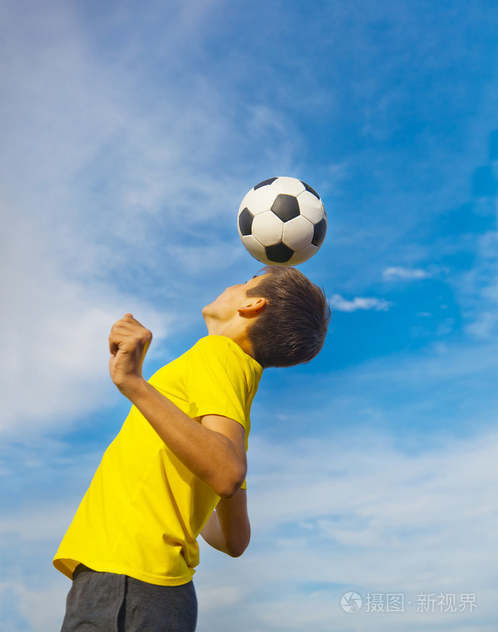 快乐的十几岁的男孩用头顶着蓝色足球球天空bac
