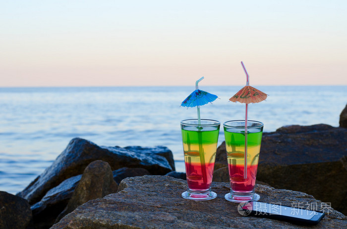 两杯鸡尾酒在海滩石头上放吸管和智能手机