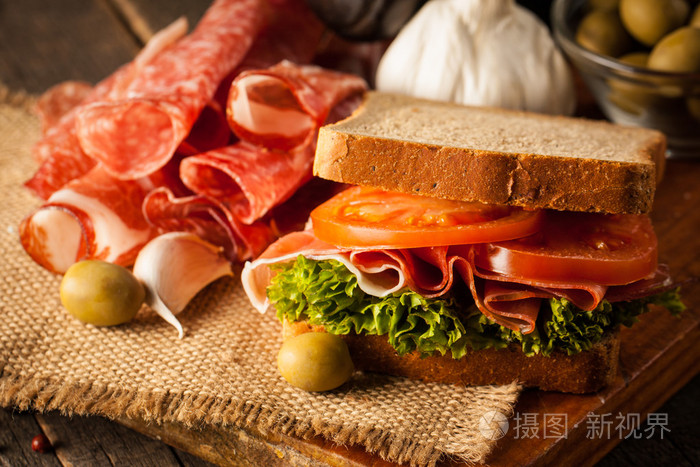 特写一张俱乐部三明治的照片。 三明治与满足意大利火腿，沙拉，蔬菜，生菜，番茄，洋葱和芥末在一个新鲜切片黑麦面包上的木制背景。 橄