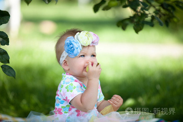 可爱的小宝贝女孩吃苹果下树在绿色的草地,在阳光明媚的美丽花园背景