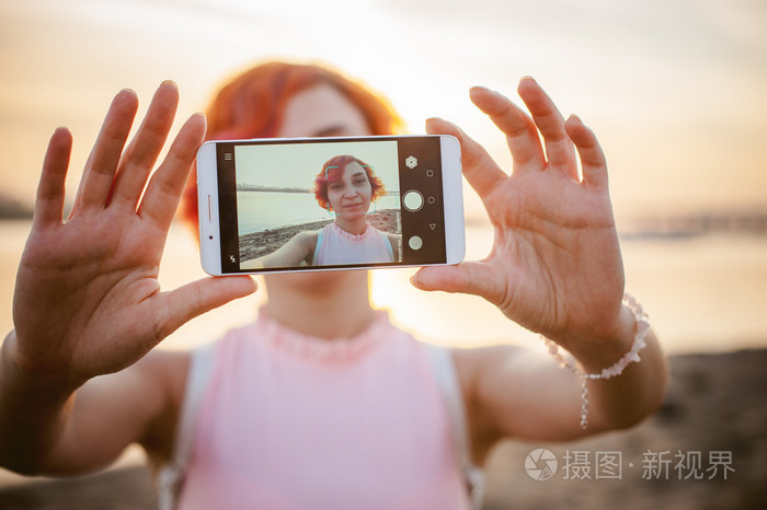 身穿浅粉色礼服红头发背包沿着河岸行走的女孩, 在温暖的夏日停泊的船只的背景下, 用手机拍摄自己的照片