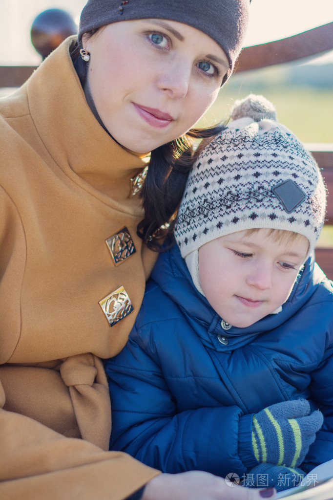 年轻女人和小男孩坐在板凳上,在秋天公园和看书.家人在一起享受时光.