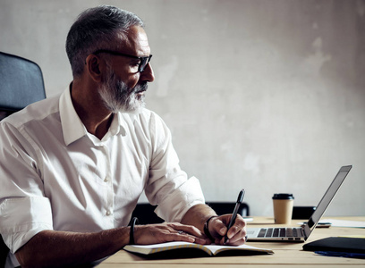成人成功的商人戴着经典的眼镜和带便携式计算机在现代阁楼木桌旁工作