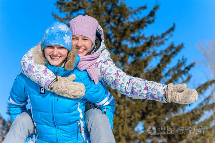 室外的肖像。两个年轻快乐的女孩玩雪在冬天的雪。骑在对方的背拥抱