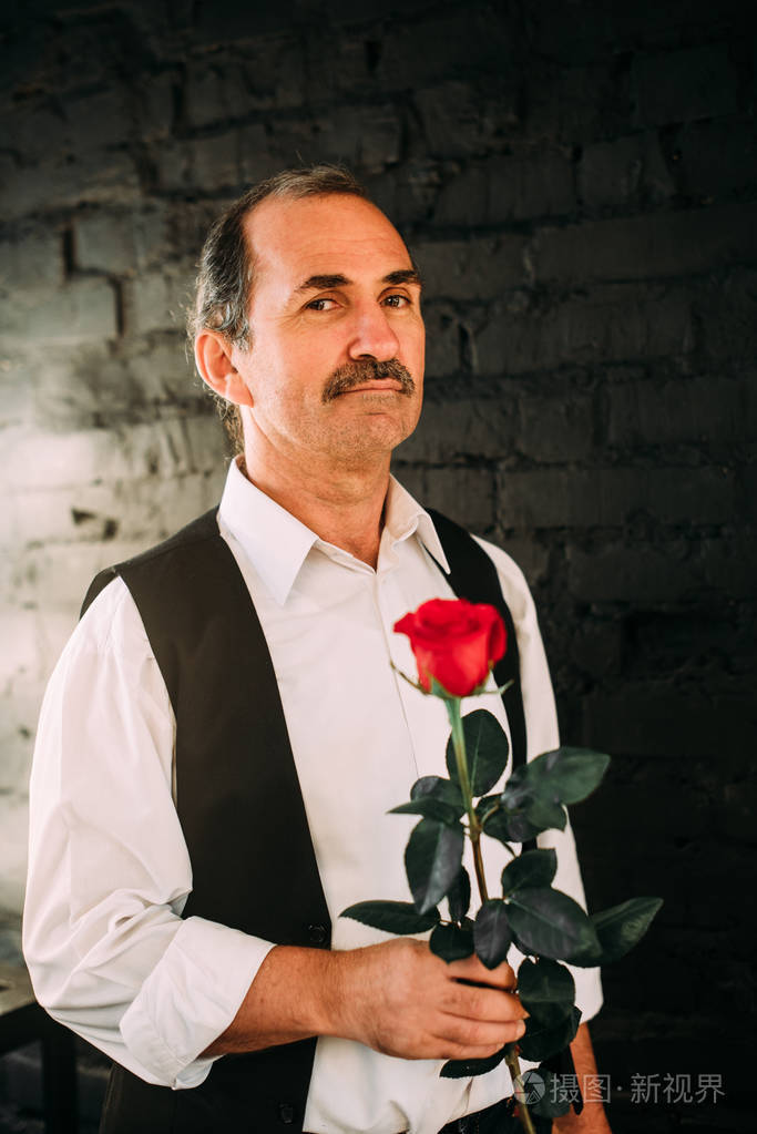 英俊的中间位老人穿着一件白衬衫和黑色的背心拿着红色的玫瑰花学士