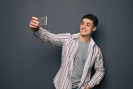 活泼开朗的年轻人在拿着手机和制作自己的照片,同时站在灰色背景下的