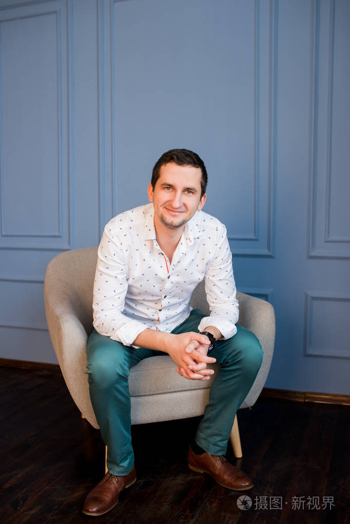 快乐英俊的男人穿着白衬衣绿色的裤子坐在扶手椅上看着相机