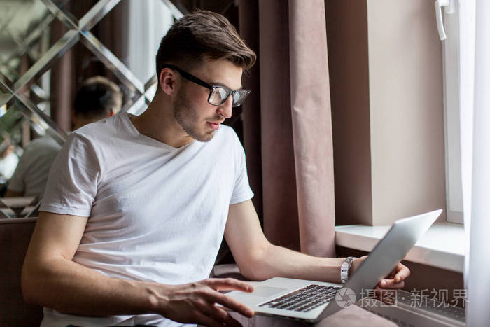英俊的大胡子的男人坐在沙发上和使用一台笔记本电脑.戴眼镜的男人