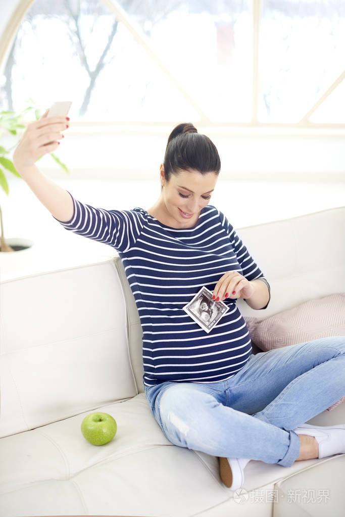 幸福的孕妇手握她的3d超声图像并在家里坐在沙发上自拍