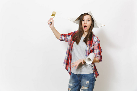 年轻震惊的美女穿着休闲服和报纸帽子,手里拿着油漆刷和壁纸卷,孤立在