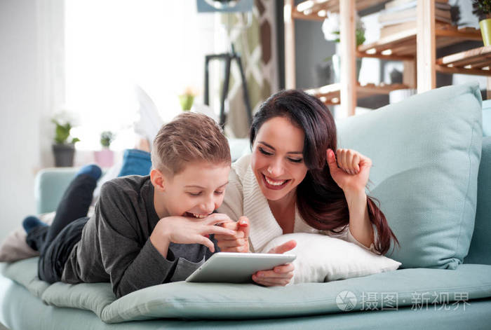 母亲和儿子躺在沙发上在家里使用数字平板电脑