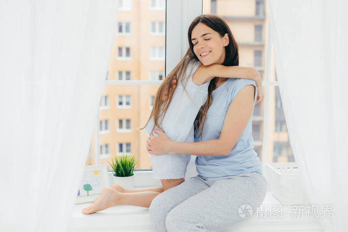 积极多情的女性和愉快的外表穿休闲的家庭服装拥抱她的小女儿表达爱