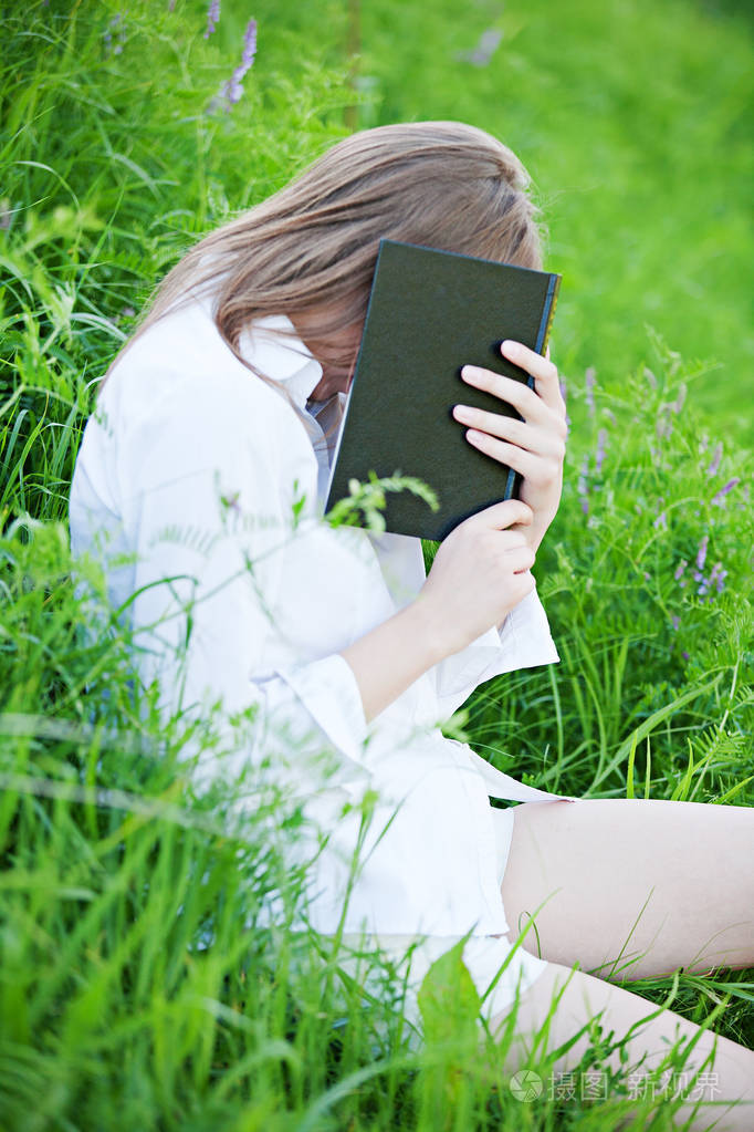 年轻漂亮的女孩坐在草地上,穿着一件白色衬衫,用一本书遮住她的脸